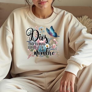 May include: A beige sweatshirt with a floral design and a butterfly. The text "Dios tiene un plan que lleva tu nombre" is printed on the sweatshirt.