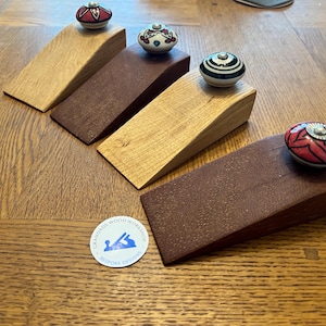 May include: Four wooden doorstops with different coloured ceramic knobs. The knobs have various patterns and designs. The doorstops are made of different types of wood, including a dark reddish brown wood and a light brown wood. The doorstops are arranged on a wooden surface.