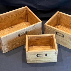 May include: Three wooden storage boxes in different sizes. The boxes are made of light-coloured wood with visible grain and have metal label holders and handles. They are arranged on a dark surface.