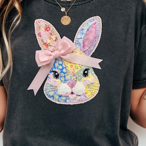 Peut inclure: T-shirt gris foncé avec un visage de lapin en patchwork et un nœud rose. Le lapin a des oreilles à imprimé floral et un motif patchwork jaune, bleu et rose. Le t-shirt est de style décontracté.
