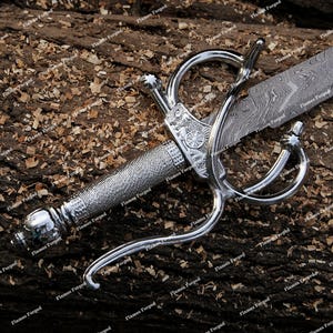 May include: A silver-toned rapier with a complex, curved metal hilt and a textured grip. The blade features a distinct wavy, layered pattern characteristic of Damascus steel. The guard includes a decorative circular emblem detail.