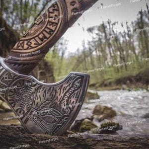 May include: A detailed, hand-carved axe with a silver head and a wooden handle. The handle features intricate carvings, and the axe head displays a decorative pattern. The axe is set against a blurred background of a stream and forest.