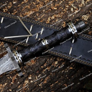 May include: A dagger with a patterned Damascus steel blade and a dark, textured wooden handle featuring metallic accents with a Greek key design. The dagger is accompanied by a black leather sheath with light-colored stitching and lacing.