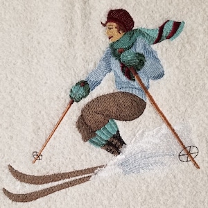 Può includere: Immagine ricamata di uno sciatore in una scena invernale. Lo sciatore indossa un maglione blu, pantaloni marroni e una sciarpa e guanti turchesi. Gli sci e i bastoncini sono marroni. Lo sfondo è bianco, a suggerire la neve.