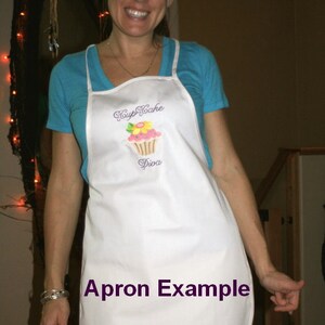 Pu&ograve; includere: Un grembiule bianco con un design di cupcake ricamato in viola e il testo "Cupcake Diva". Il grembiule &egrave; indossato da una persona con una camicia blu. Il testo "Apron Example" &egrave; scritto in viola sul grembiule.