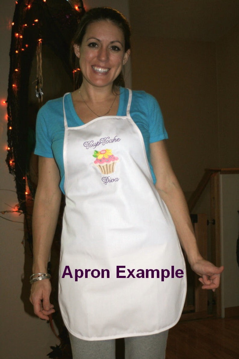 Pu&ograve; includere: Un grembiule bianco con un design di cupcake ricamato in viola e il testo "Cupcake Diva". Il grembiule &egrave; indossato da una persona con una camicia blu. Il testo "Apron Example" &egrave; scritto in viola sul grembiule.