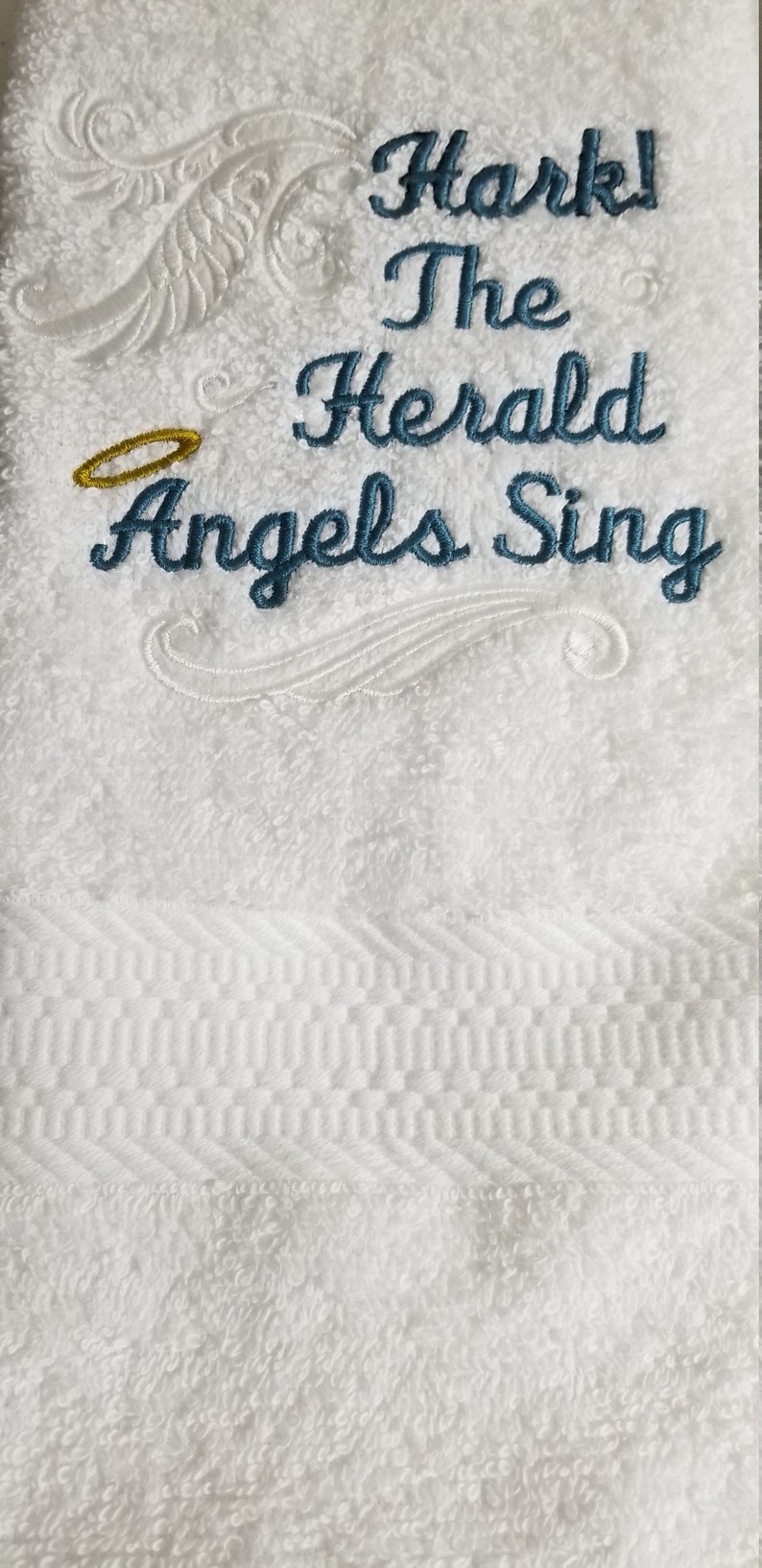 Pu&ograve; includere: Asciugamano bianco con ricamo blu che recita "Hark! The Herald Angels Sing". Un'aureola dorata &egrave; ricamata sopra il testo.