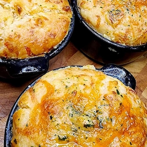 Könnte beinhalten: Vier einzelne Portionen käsigen, goldbraunen Auflaufs in schwarzen gusseisernen Auflaufformen. Der Auflauf ist mit Kräutern und einer geschmolzenen Käsesauce überzogen.