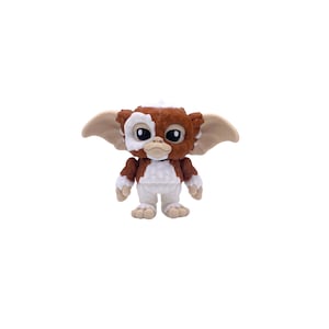 May include: A small, detailed figurine of a Gremlin. The figure has a brown and white head with large black eyes, tan ears, and a white patch over one eye. The body is white with brown accents, and the figure is standing.