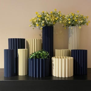 以下が含まれることがあります： クリーム色とネイビーブルーの2色でできた、装飾的な花瓶が9つあります。花瓶はすべて円筒形で、リブ状のテクスチャがあります。花瓶の中には、黄色い花が飾られているものもあります。