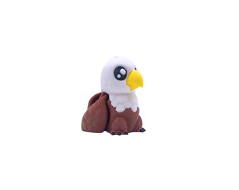 May include: A small, stylized bald eagle figurine. The eagle has a white head and chest, brown wings, and a yellow beak. Its large eyes are black. The figurine is sitting down and has small black feet.