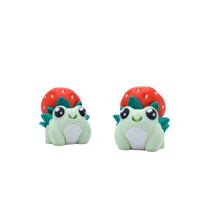 May include: Two charming frog figurines in light green, each with a white belly and large eyes. They are paired with a red strawberry with green leaves. These decorative figurines are likely handmade and suitable for display.