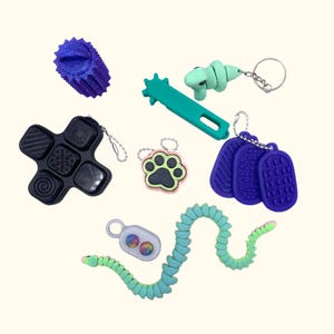 Könnte beinhalten: Eine Sammlung farbenfroher Zappelspielzeuge in verschiedenen Formen. Enthält einen blauen Stachelball, eine grüne Schlange, ein schwarzes kreuzförmiges Spielzeug, einen grünen Dinosaurier-Schlüsselanhänger, einen Pfotenabdruck-Schlüsselanhänger und drei blaue strukturierte Anhänger.