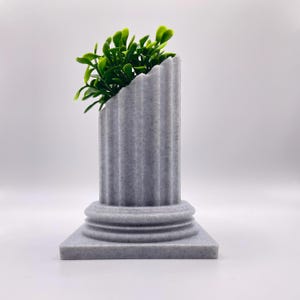 Puede incluir: Un macetero de hormigón gris con forma de columna con una planta verde que crece en la parte superior.