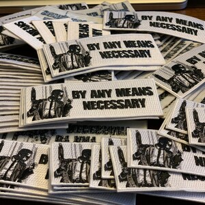 May include: A pile of white fabric patches with black embroidery. The patches feature a man in a suit with the text "By Any Means Necessary".