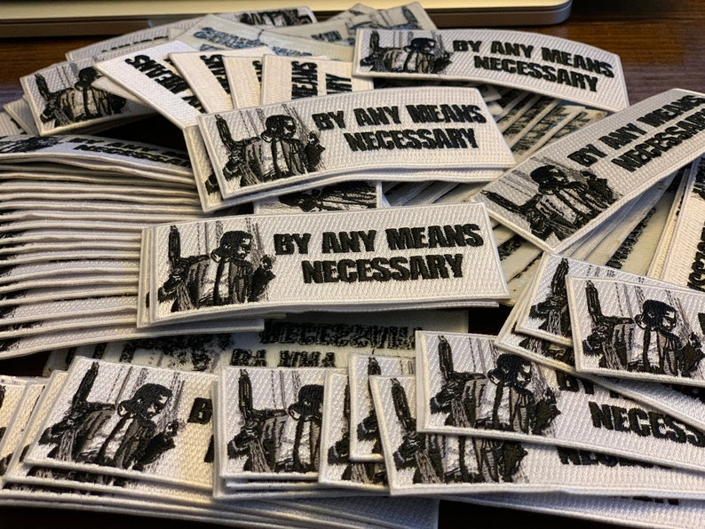 May include: A pile of white fabric patches with black embroidery. The patches feature a man in a suit with the text "By Any Means Necessary".