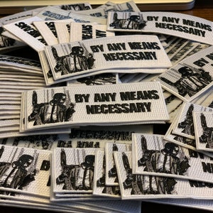 May include: A pile of white fabric patches with black embroidery. The patches feature a man in a suit with the text "By Any Means Necessary".