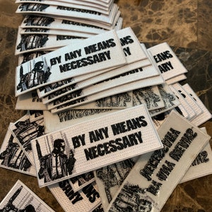 May include: A stack of white embroidered patches with the text "By Any Means Necessary" and a black and white image of a man in a suit.