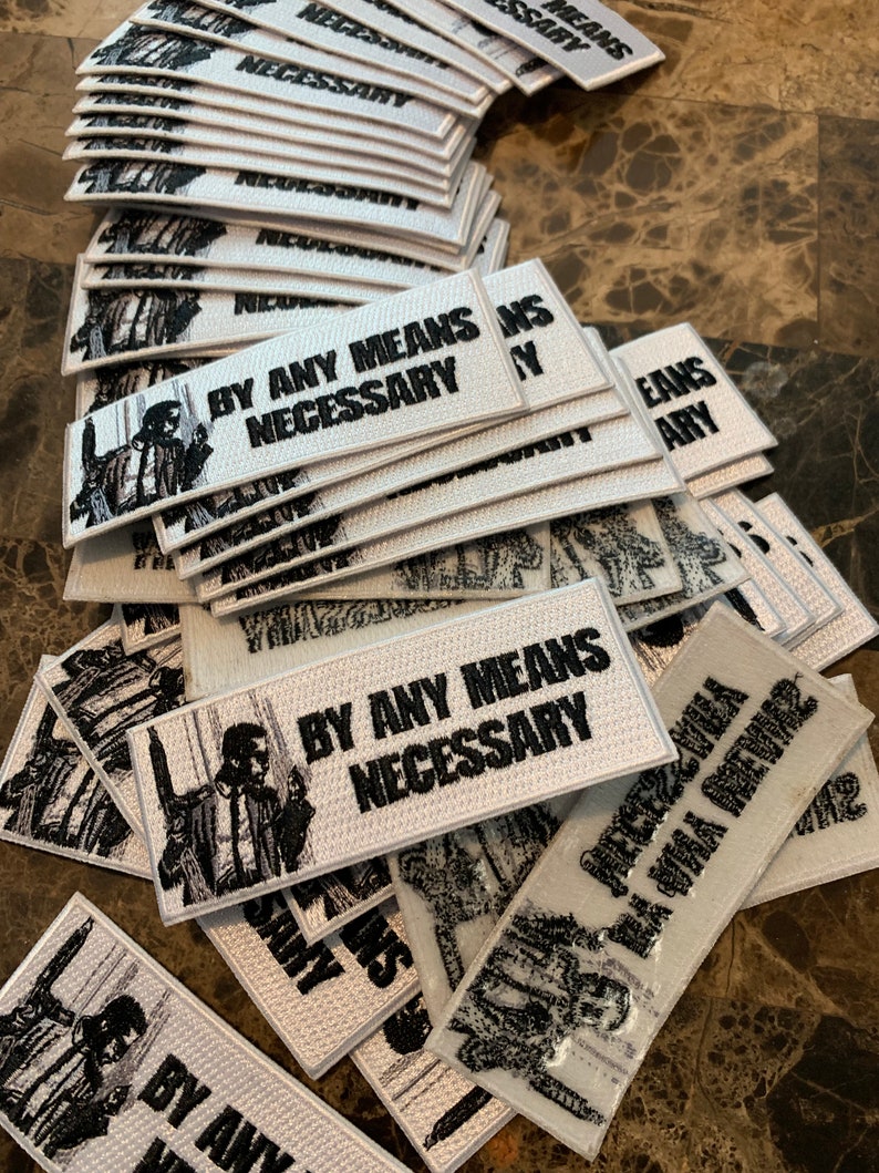 May include: A stack of white embroidered patches with the text "By Any Means Necessary" and a black and white image of a man in a suit.