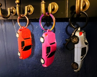 May include: Three car-shaped keychains in red, pink, and white hang from a brass hook. The keychains have black accents and silver-colored wheels. The keyrings are attached to the car keychains with a metal ring.
