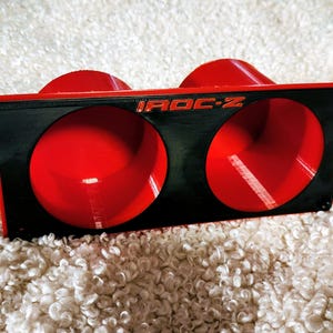 May include: A black and red automotive part with the text "IROC-Z" in red. The part has two circular openings and a triangular shape on each end. The interior of the openings is red.