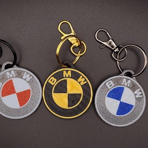 May include: Three BMW logo keychains. One has a red and white design, another has a black and yellow design, and the last has a blue and white design. Each keychain has a metal clasp and ring.