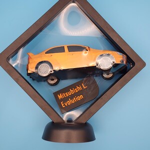 May include: An orange model car, a Mitsubishi L. Evolution, is displayed in a black, diamond-shaped frame with a clear cover. The car has silver wheels and a rear spoiler. The frame sits on a black base against a blue background.