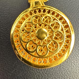 K&ouml;nnte beinhalten: Eine goldfarbene Halskette mit Anh&auml;nger und einem aufwendigen, durchbrochenen Design. Der kreisf&ouml;rmige Anh&auml;nger hat einen zentralen, erhabenen Kreis, der von einem Blumenmuster und detailliertem Filigran umgeben ist. Die Kette ist ebenfalls goldfarben.