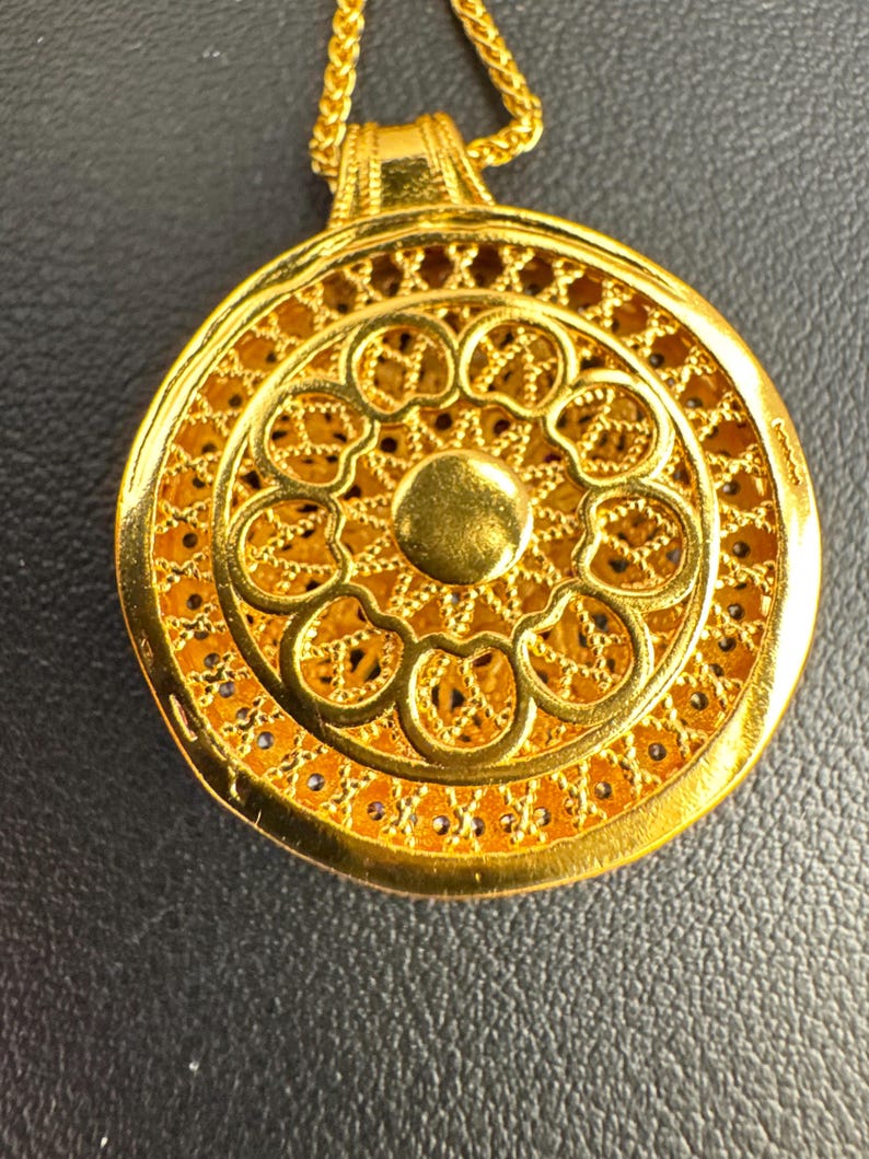 K&ouml;nnte beinhalten: Eine goldfarbene Halskette mit Anh&auml;nger und einem aufwendigen, durchbrochenen Design. Der kreisf&ouml;rmige Anh&auml;nger hat einen zentralen, erhabenen Kreis, der von einem Blumenmuster und detailliertem Filigran umgeben ist. Die Kette ist ebenfalls goldfarben.