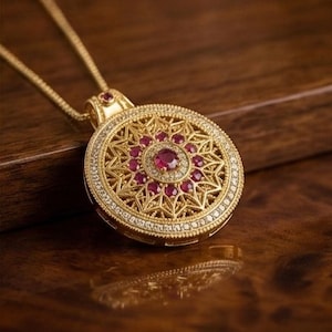 K&ouml;nnte beinhalten: Eine goldfarbene Halskette mit Anh&auml;nger und kreisf&ouml;rmigem Design. Der Anh&auml;nger hat einen zentralen Rubin, der von kleineren Rubinen und klaren Edelsteinen umgeben ist. Der &auml;u&szlig;ere Rand ist ebenfalls mit klaren Edelsteinen besetzt. Die Halskette liegt auf einer Holzoberfl&auml;che.