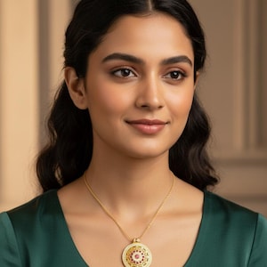 May include: A gold-colored pendant necklace featuring a circular pendant with a central red gemstone surrounded by smaller red and white stones. The pendant has an ornate, detailed design and hangs from a delicate gold chain. The woman is wearing a green top.