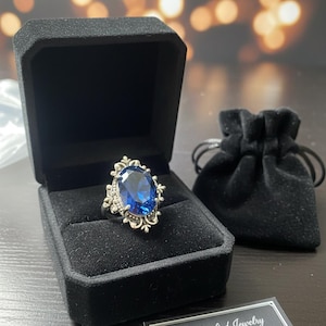 May include: A silver ring with a large, oval, deep blue gemstone, set in an ornate filigree design. The ring is displayed in a black velvet box, with a small black velvet pouch and a tag that reads "Handcrafted Jewelry Deep Blue Dragon Ring."