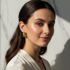 May include: A woman with dark hair wearing gold hoop earrings. The earrings have a detailed design and are a bright gold colour. She is wearing a white top. The background is a white marble wall.