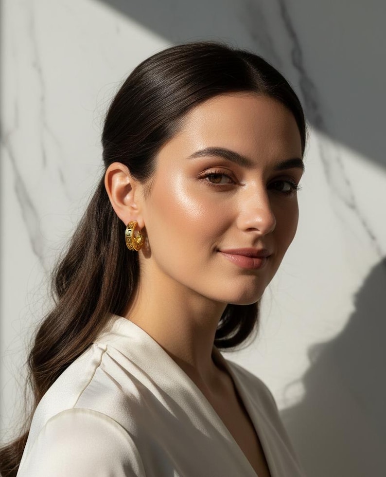 May include: A woman with dark hair wearing gold hoop earrings. The earrings have a detailed design and are a bright gold colour. She is wearing a white top. The background is a white marble wall.