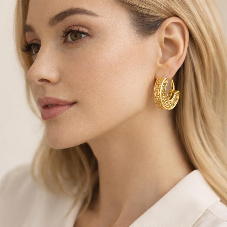May include: Gold-coloured hoop earrings featuring a Greek key design. The earrings are a half-circle shape with a detailed pattern. The earrings are worn on the ear.