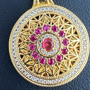 K&ouml;nnte beinhalten: Goldfarbener Anh&auml;nger mit kreisf&ouml;rmigem Design. Der Anh&auml;nger hat einen zentralen Rubin, der von kleineren Rubinen und klaren Edelsteinen umgeben ist. Der &auml;u&szlig;ere Rand ist mit klaren Edelsteinen besetzt, und der Anh&auml;nger h&auml;ngt an einer goldenen Kette.