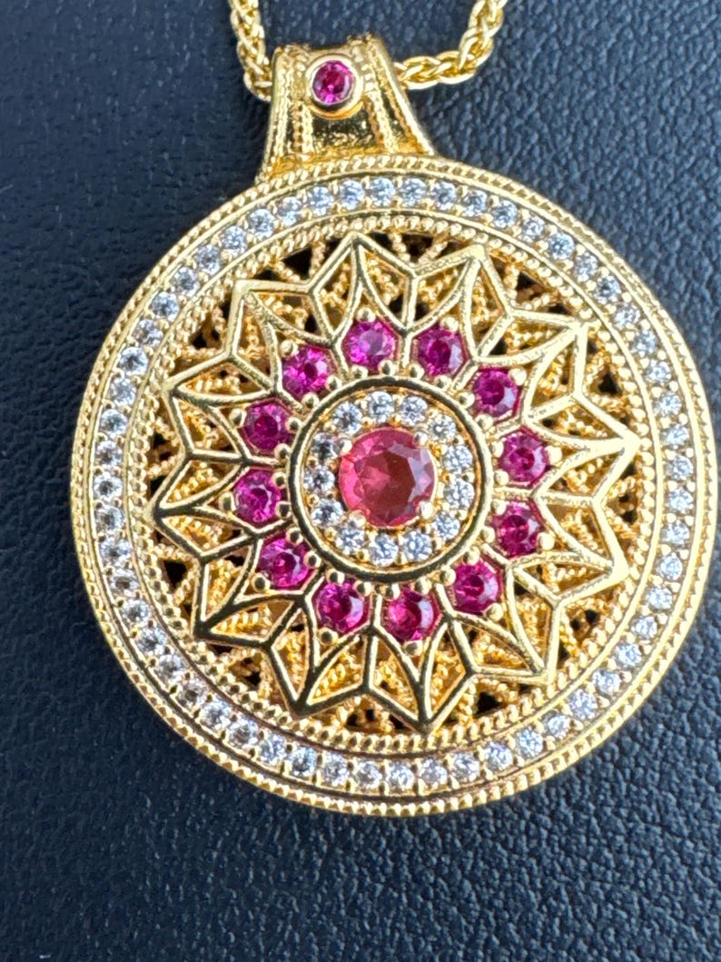 K&ouml;nnte beinhalten: Goldfarbener Anh&auml;nger mit kreisf&ouml;rmigem Design. Der Anh&auml;nger hat einen zentralen Rubin, der von kleineren Rubinen und klaren Edelsteinen umgeben ist. Der &auml;u&szlig;ere Rand ist mit klaren Edelsteinen besetzt, und der Anh&auml;nger h&auml;ngt an einer goldenen Kette.