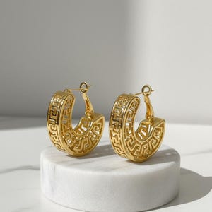 May include: A pair of gold hoop earrings featuring a Greek key pattern. The earrings are presented on a white marble surface against a neutral backdrop. The earrings have a hinged clasp.