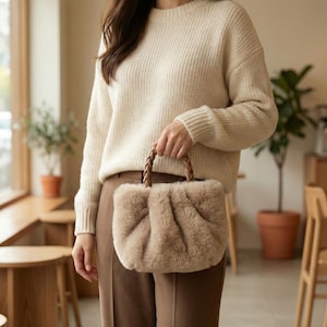May include: A beige faux fur handbag with a plaited brown handle. The bag has a gathered design and is held by a person wearing a cream-coloured jumper and brown trousers. The background includes wooden furniture and potted plants.