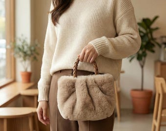 Könnte beinhalten: Eine beige Kunstfell-Handtasche mit einem geflochtenen braunen Griff. Die Tasche hat ein gerafftes Design und wird von einer Person gehalten, die einen cremefarbenen Pullover und eine braune Hose trägt. Der Hintergrund umfasst Holzmöbel und Topfpflanzen.