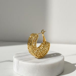 May include: A gold-coloured hoop earring featuring a Greek key pattern. The earring has a thick, rounded design with the key pattern carved into the surface. It is displayed on a white, round pedestal.