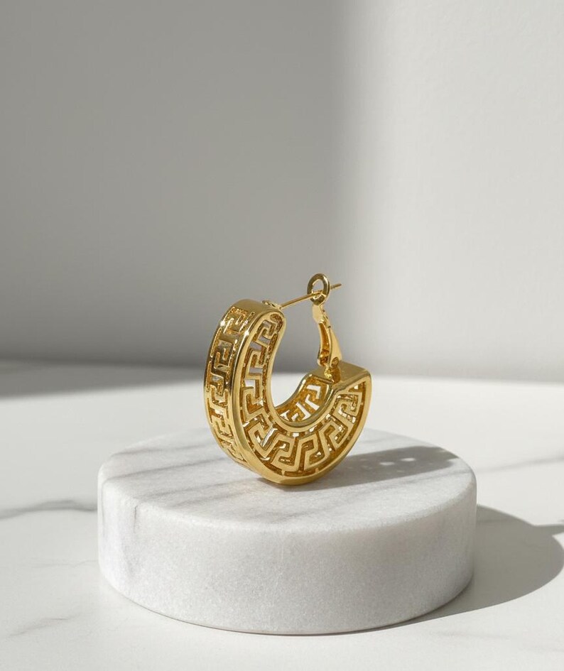 May include: A gold-coloured hoop earring featuring a Greek key pattern. The earring has a thick, rounded design with the key pattern carved into the surface. It is displayed on a white, round pedestal.