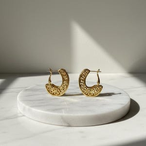 May include: A pair of gold-coloured hoop earrings with a Greek key design. The earrings are crescent-shaped and feature a repeating geometric pattern. They are displayed on a white marble surface.