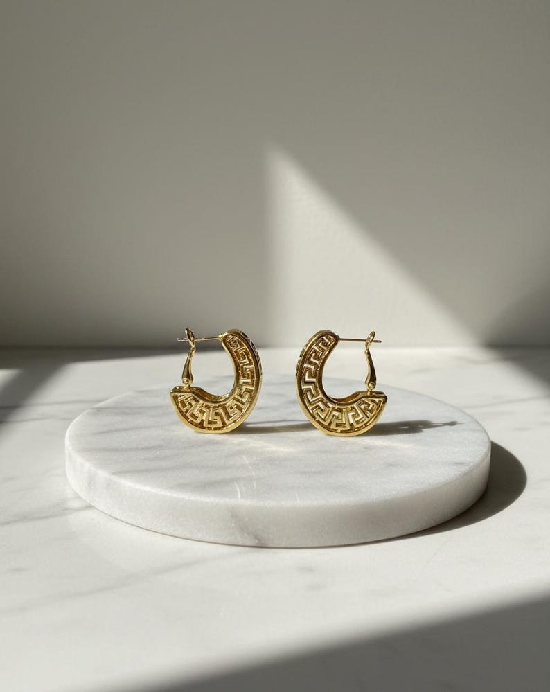 May include: A pair of gold-coloured hoop earrings with a Greek key design. The earrings are crescent-shaped and feature a repeating geometric pattern. They are displayed on a white marble surface.