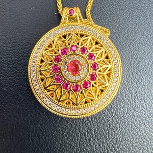 K&ouml;nnte beinhalten: Eine goldfarbene Anh&auml;ngerhalskette mit kreisf&ouml;rmigem Design. Der Anh&auml;nger hat einen zentralen roten Edelstein, der von kleineren roten Edelsteinen und klaren Kristallen umgeben ist. Die Halskette hat eine zarte Goldkette.