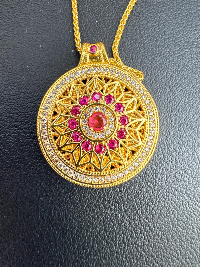 K&ouml;nnte beinhalten: Eine goldfarbene Anh&auml;ngerhalskette mit kreisf&ouml;rmigem Design. Der Anh&auml;nger hat einen zentralen roten Edelstein, der von kleineren roten Edelsteinen und klaren Kristallen umgeben ist. Die Halskette hat eine zarte Goldkette.