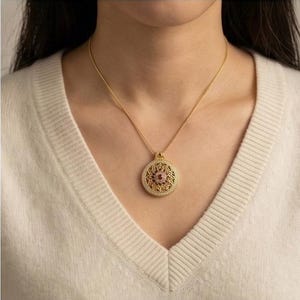 K&ouml;nnte beinhalten: Eine goldfarbene Halskette mit Anh&auml;nger und einem runden, verzierten Anh&auml;nger. Der Anh&auml;nger hat einen zentralen rosa Edelstein, umgeben von einem detaillierten, bl&uuml;tenartigen Design und kleinen, klaren Steinen. Die Halskette wird von einer Person getragen, die einen cremefarbenen Pullover tr&auml;gt.