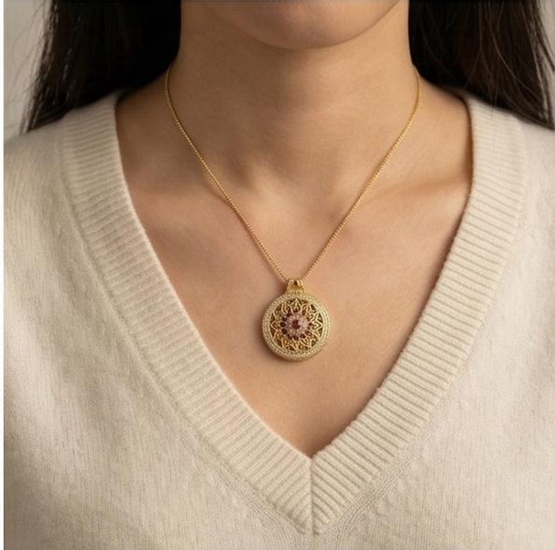 K&ouml;nnte beinhalten: Eine goldfarbene Halskette mit Anh&auml;nger und einem runden, verzierten Anh&auml;nger. Der Anh&auml;nger hat einen zentralen rosa Edelstein, umgeben von einem detaillierten, bl&uuml;tenartigen Design und kleinen, klaren Steinen. Die Halskette wird von einer Person getragen, die einen cremefarbenen Pullover tr&auml;gt.