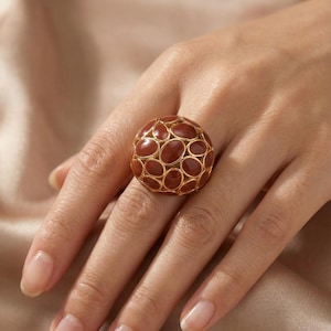May include: A statement ring with a spherical design. The ring has a gold-coloured metal frame with oval, reddish-brown stones. The ring is worn on a hand with manicured nails, against a neutral background. The ring's design is eye-catching and bold.