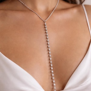 May include: A silver Y-shaped necklace featuring a fine chain and a vertical line of clear, sparkling crystals. The necklace is set against a white satin top, creating a contrast of textures and colours. The jewellery is a simple yet elegant accessory.